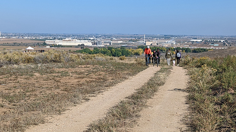 A wood trailhead sign stands on a dirt trail with wood fence and a grass prairie and blue sky.